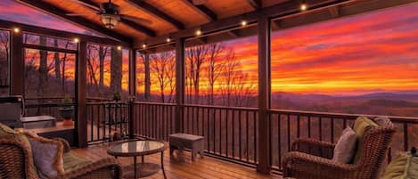 A covered deck with wicker furniture, a table, and a view of a sunset over a forested landscape.