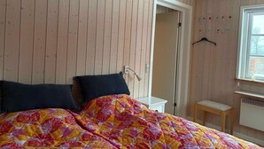 Ein Zimmer mit Holzwänden, einem Bett mit farbenfroher Blumenbettdecke, einem weißen Nachttisch, einem kleinen Holzschemel und einem Fenster mit Blick.