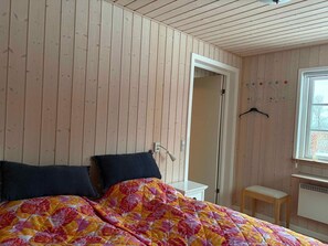 A room with wooden walls, a bed with a colorful floral bedspread, a white bedside table, a small wooden stool, and a window with a view. - 4 Star Holiday Home in Roslev (Roslev)