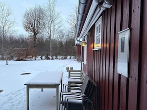 A covered outdoor seating area with a table and chairs, a red building with a window, and a snowy landscape. - 4 Star Holiday Home in Roslev (Roslev)