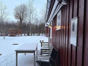 A covered outdoor seating area with a table and chairs, a red building with a window, and a snowy landscape. - 4 Star Holiday Home in Roslev (Roslev)