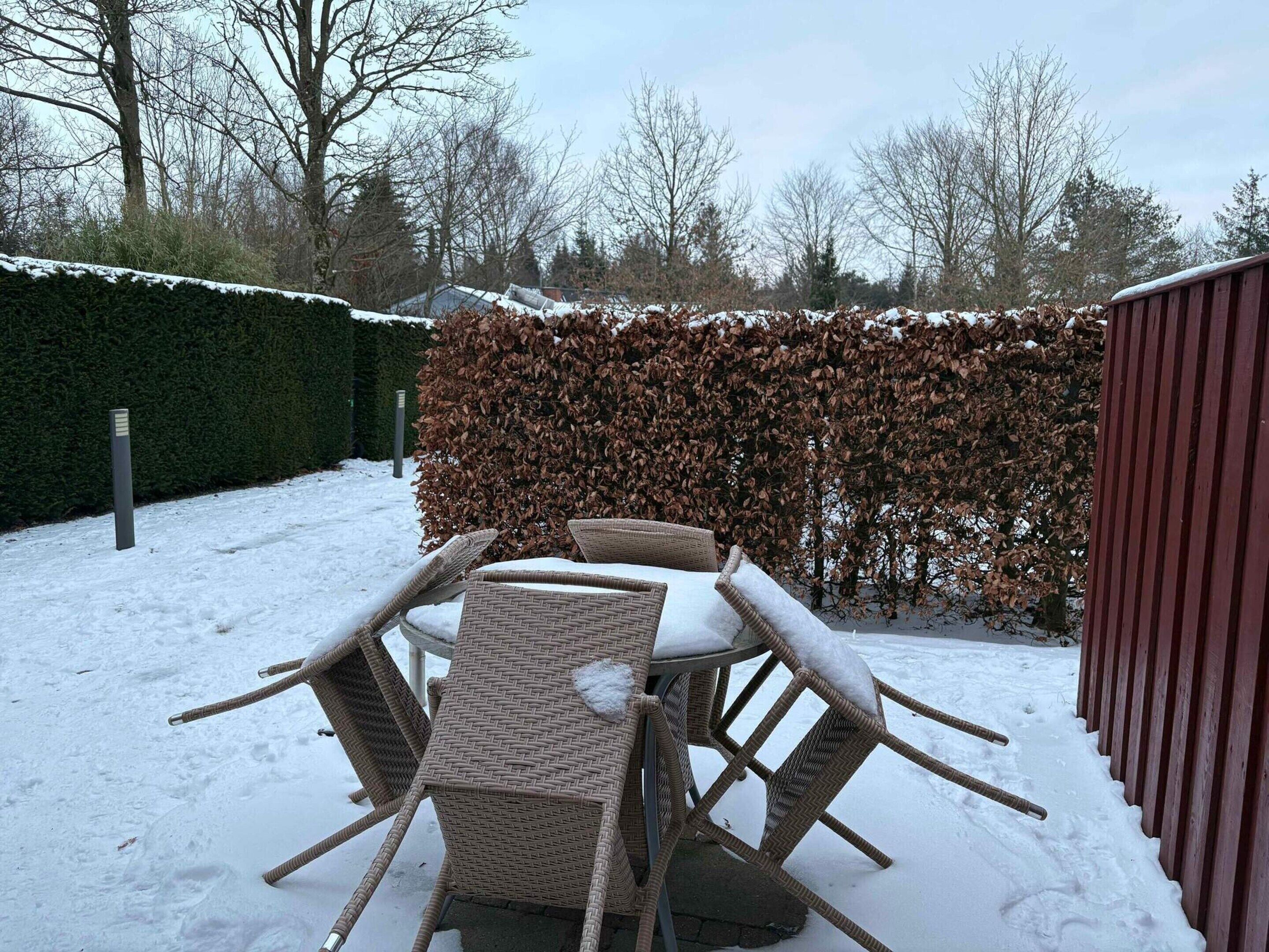 A patio with a table and chairs covered in snow, surrounded by a hedge and a wooden fence.