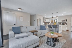 A modern living room with a grey sectional sofa, a wooden coffee table, and a kitchen in the background. - Edmonton's Ultimate Family Hangout: Retro Gaming Garage and Private Hot Tub! (Edmonton)