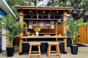 Un bar al aire libre con estructura de madera, dos taburetes, una mesa y plantas en macetas.
