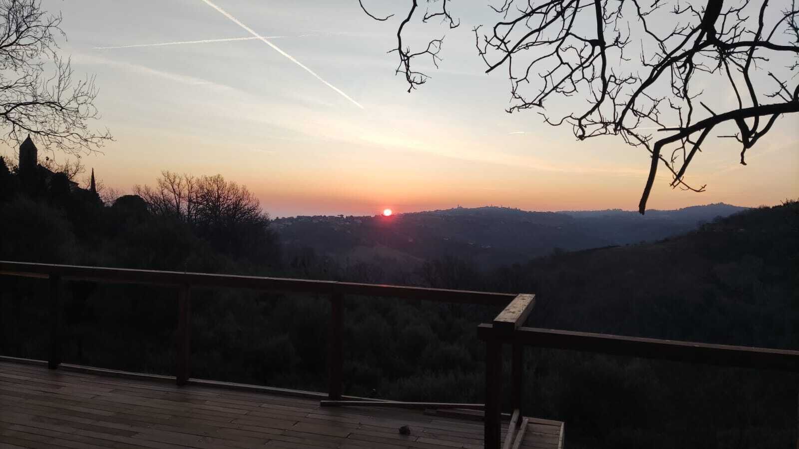 Solnedgangsudsigten fra en træterrasse over et bølgende landskab med nøgne træer og en bygning i det fjerne.