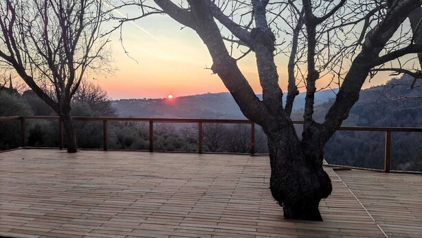 A wooden deck with a railing, a tree in the foreground, and a sunset view in the background.