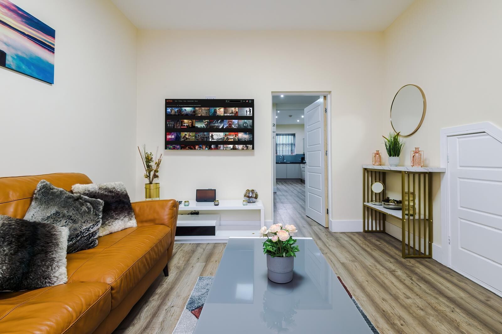 A living room with a leather sofa, a coffee table with a vase of flowers, a TV mounted on the wall displaying movie options, and a doorway leading to a kitchen.
