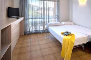 A hotel room with a bed, a TV on a wooden shelf, a window with curtains, and a yellow towel on the bed. - Apartment in Tossa de Mar Near Beach (Tossa de Mar)