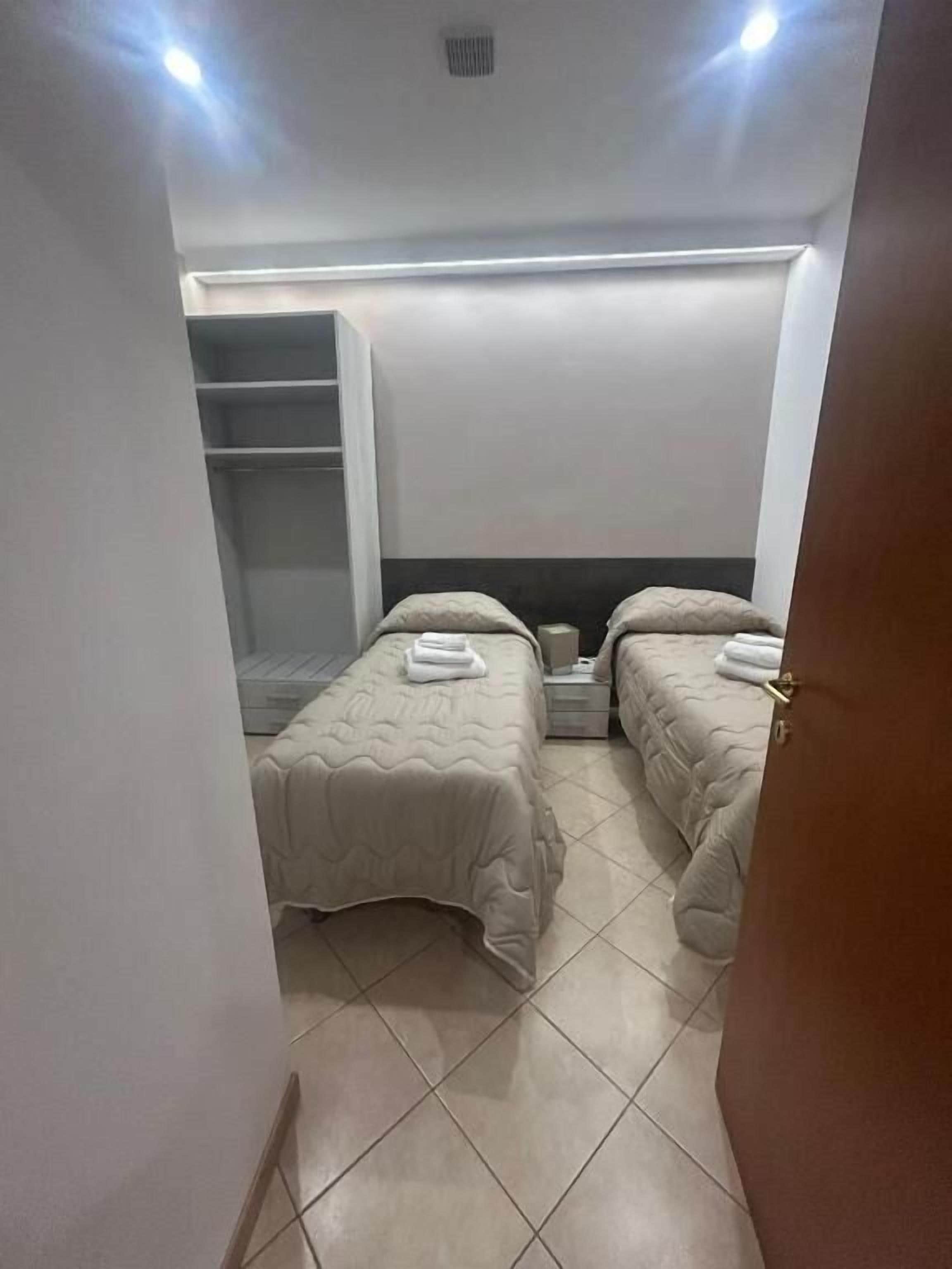 Quarto pequeno e bem iluminado, com duas camas, um guarda-roupa e uma mesa de cabeceira.