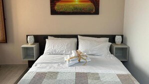 Habitación de hotel con una cama, dos mesitas de noche, un cuadro de un atardecer y un conjunto de toallas sobre la cama.