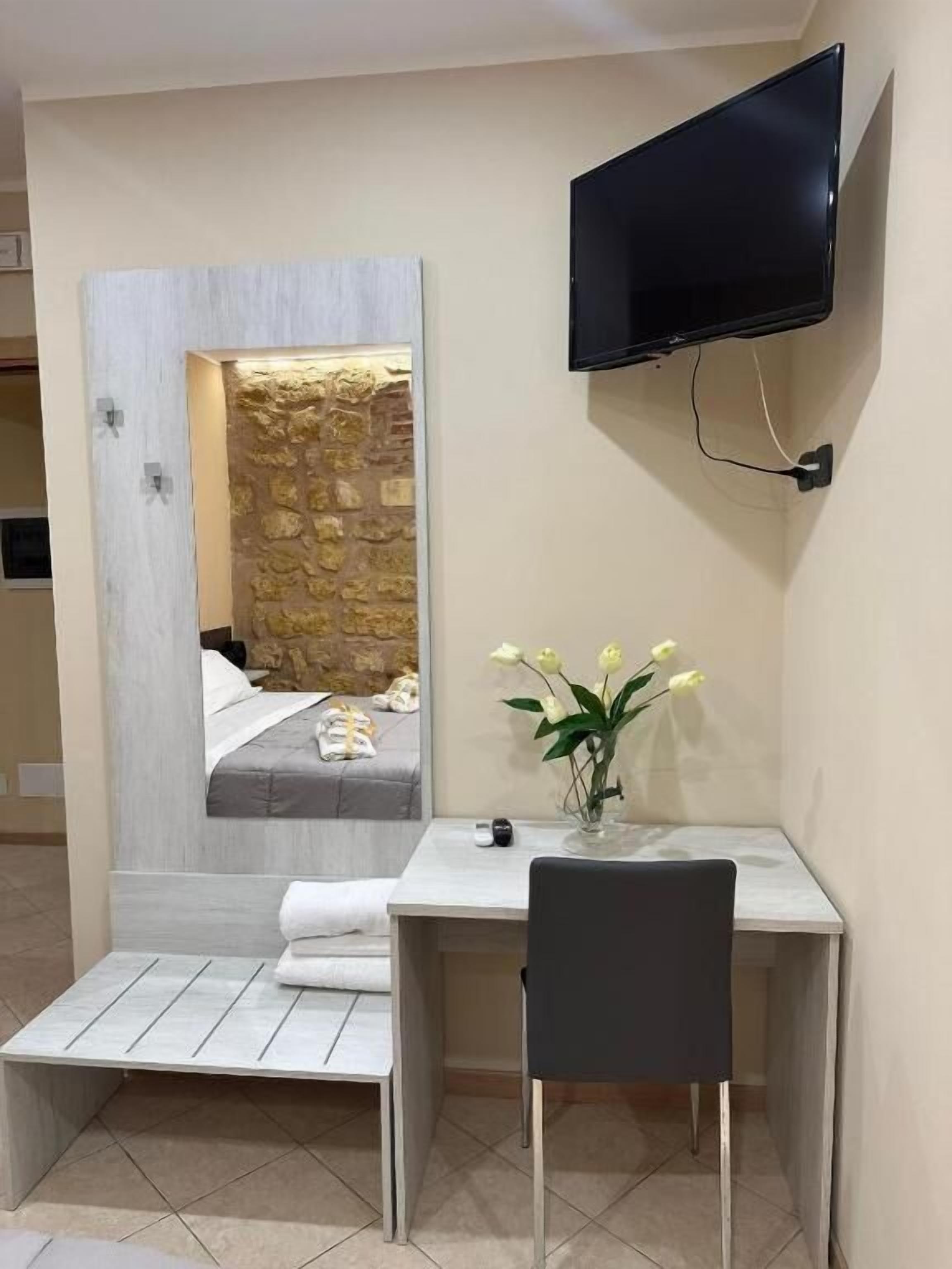 A hotel room with a bed, a TV mounted on the wall, a desk with a chair, and a vase with flowers.