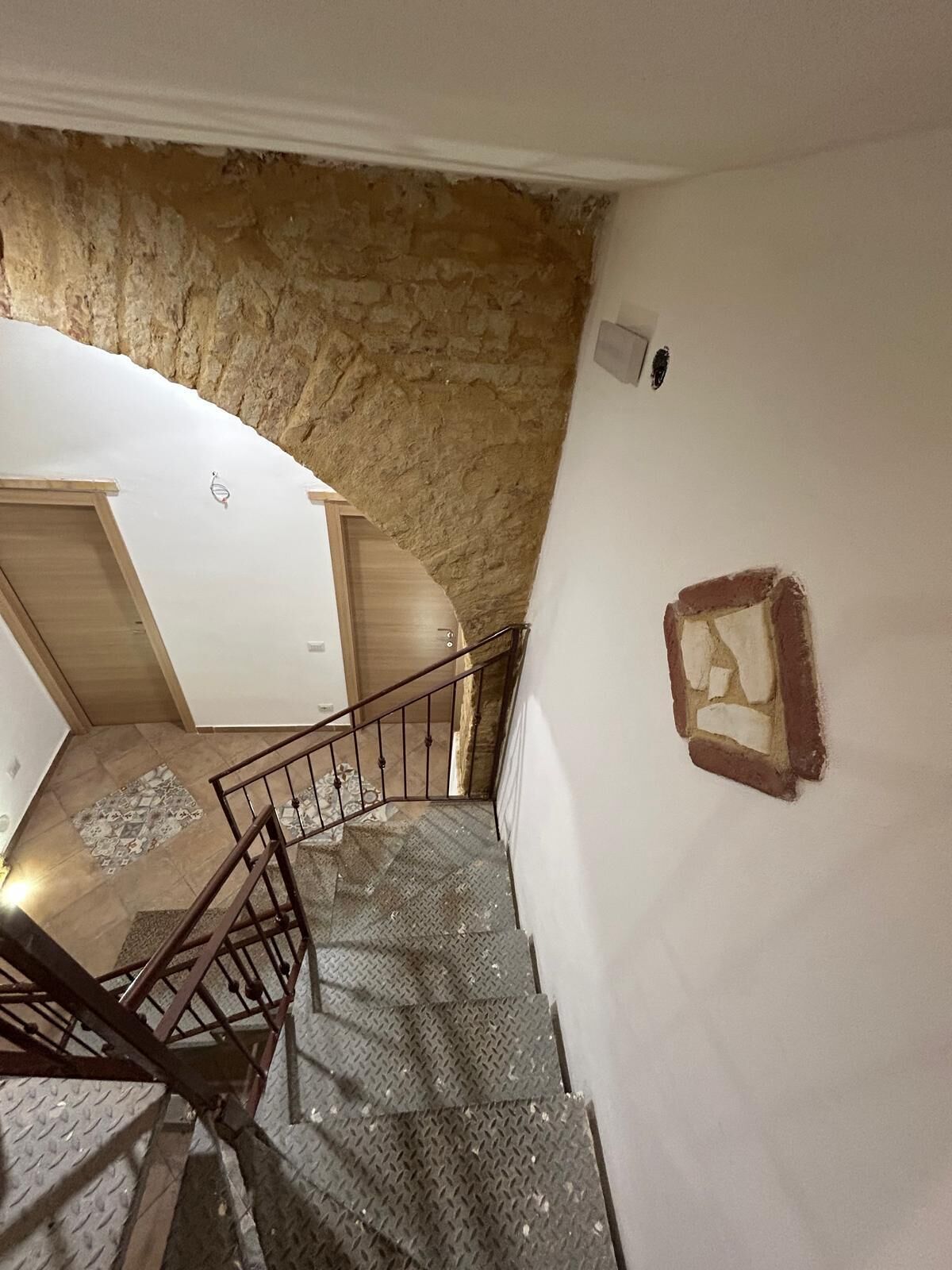 Una scala con ringhiera in metallo che conduce a una stanza con parete in pietra e una piccola cornice appesa al muro.