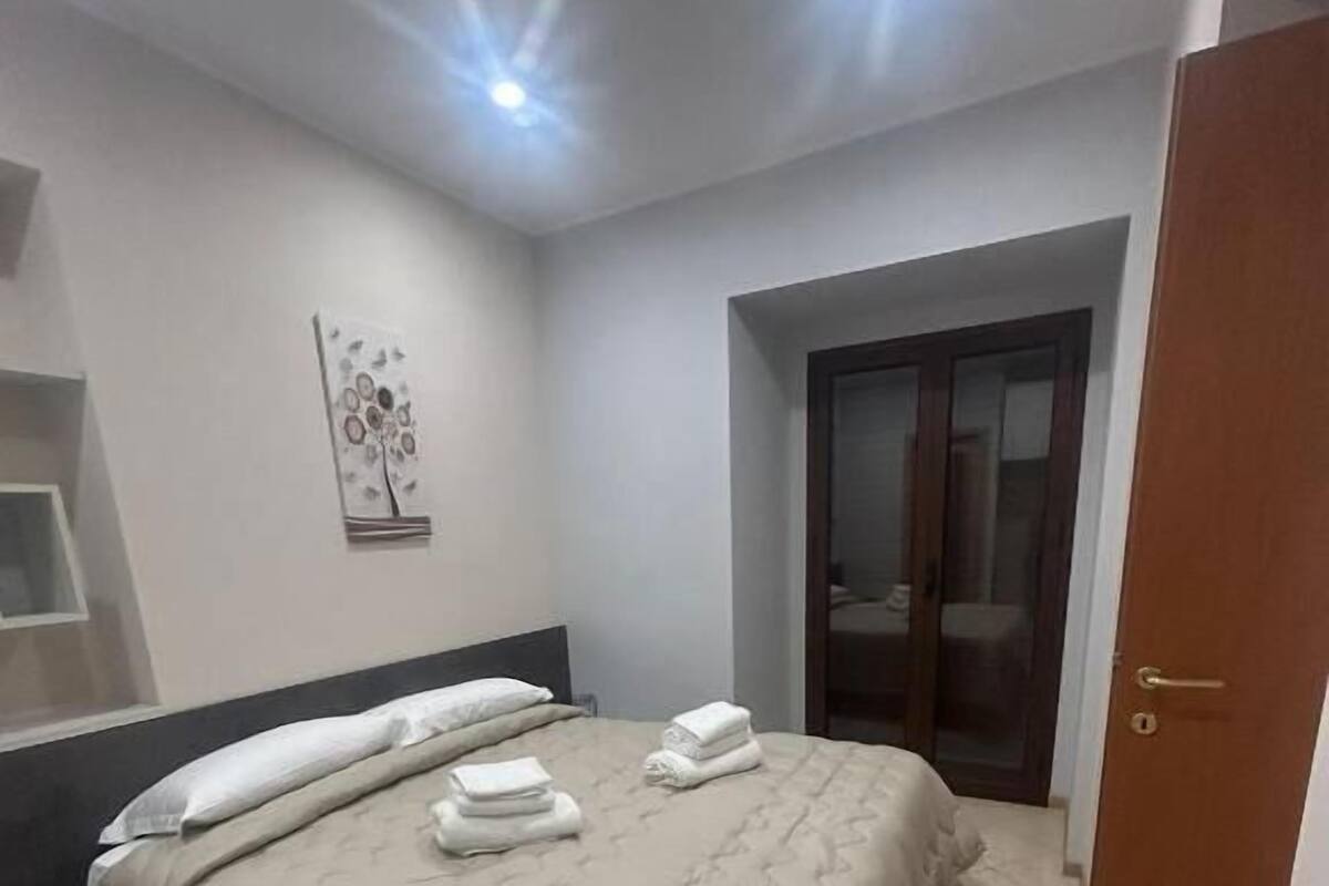 A bedroom with a bed, a nightstand, a wall-mounted artwork, a door, and a tiled floor.