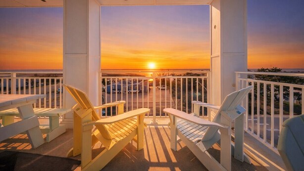 A covered outdoor area with a view of the sunset over the ocean, featuring a railing and several Adirondack chairs.