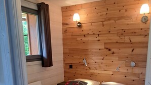 Ein Zimmer mit Holzwand, einem Bett mit gemustertem Überwurf, zwei Kissen, einem Fenster mit Vorhängen und zwei Wandleuchten.
