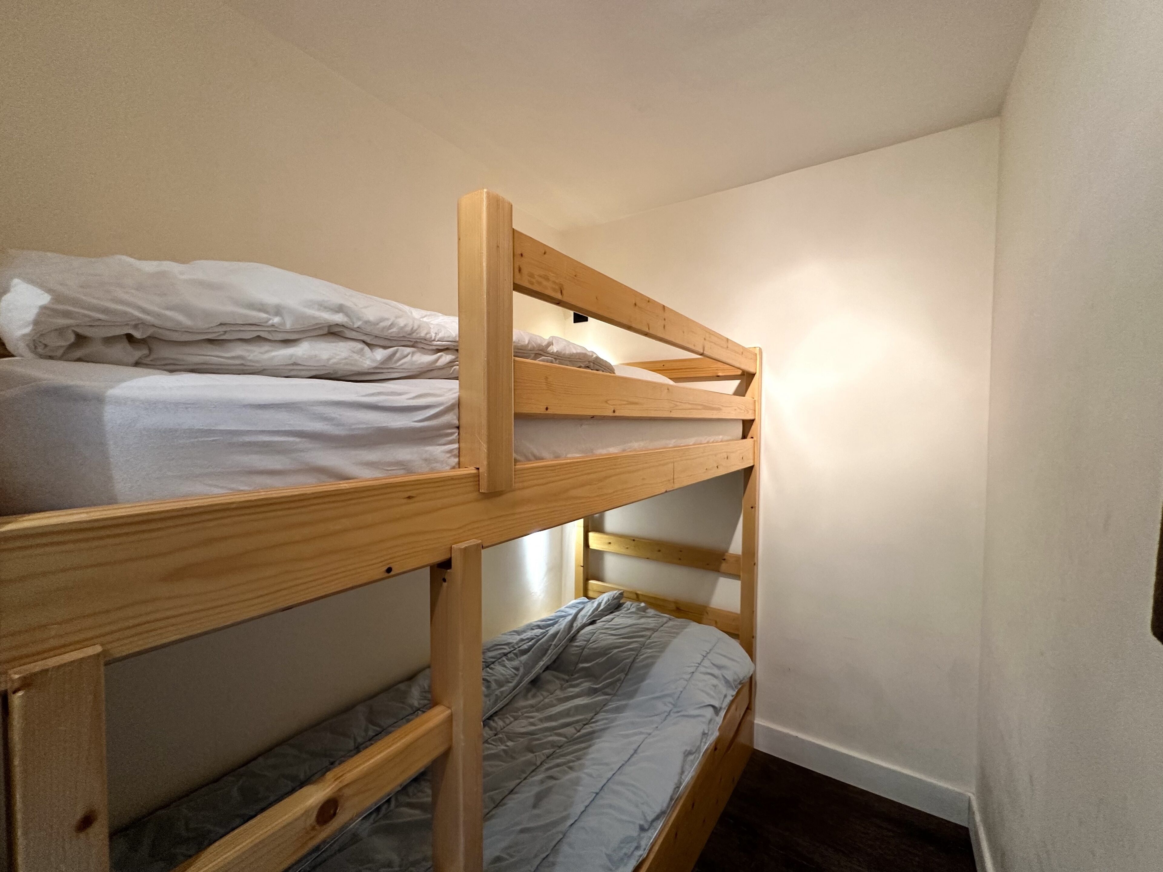 Una cama litera con estructura de madera, la cama inferior con colchón gris y la superior con ropa de cama blanca.