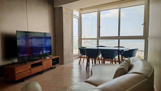 A modern living room with a large flat-screen TV, a leather sofa, a dining table with chairs, and a large window offering a view of the cityscape. - Brilliant 3-bedroom apartment with AC in vibrant Panamá (Panamá)