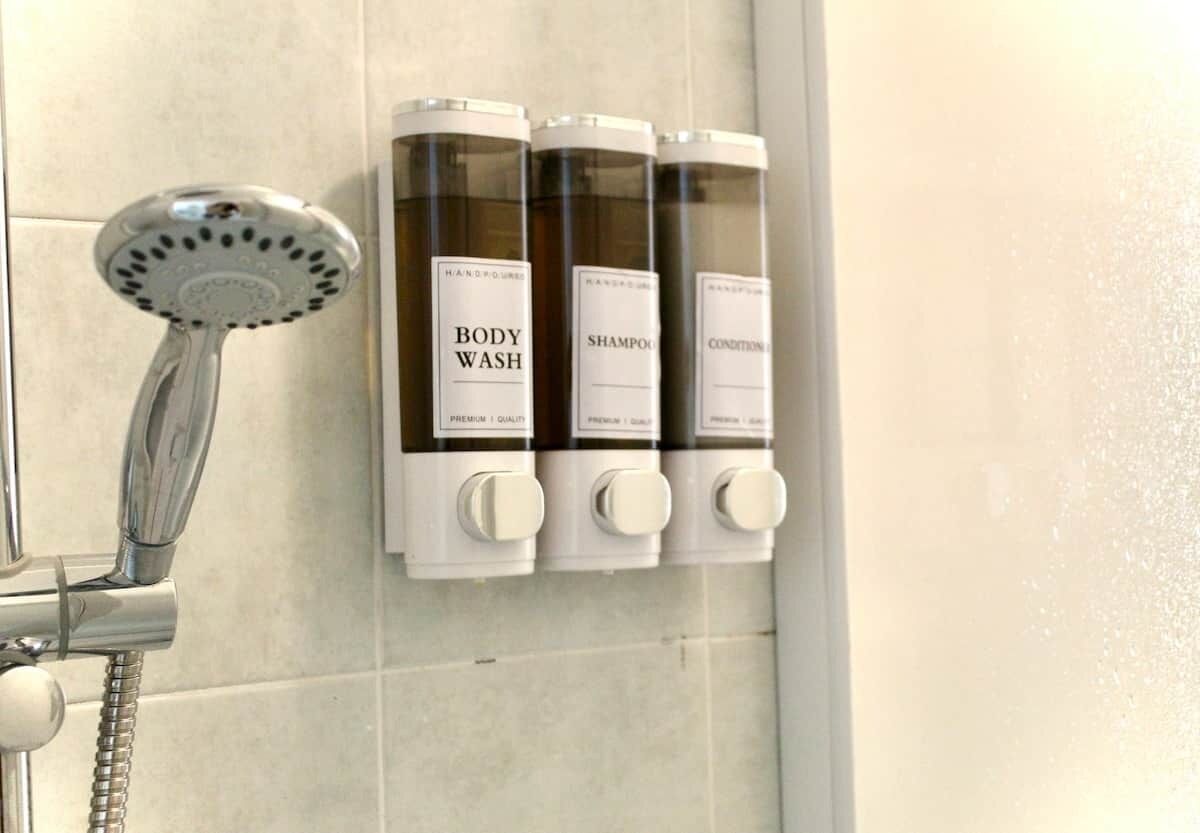 Ducha con cabezal de ducha de mano y tres botellas de gel de baño, champú y acondicionador en un dispensador montado en la pared.