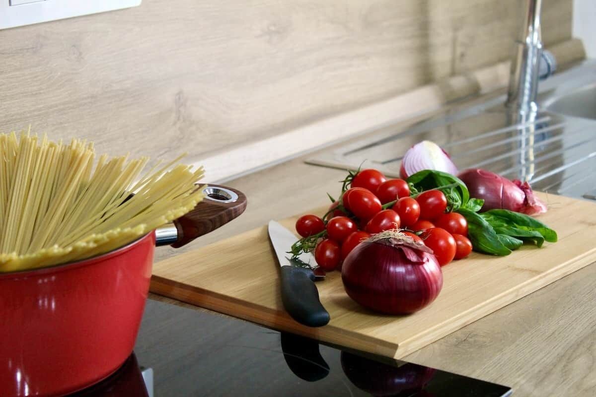 A kitchen counter with a pot of spaghetti, a cutting board with tomatoes, onions, and basil, and a knife.