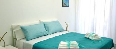 A bed with a teal bedspread and pillows, a nightstand, a framed picture on the wall, and a window with curtains.