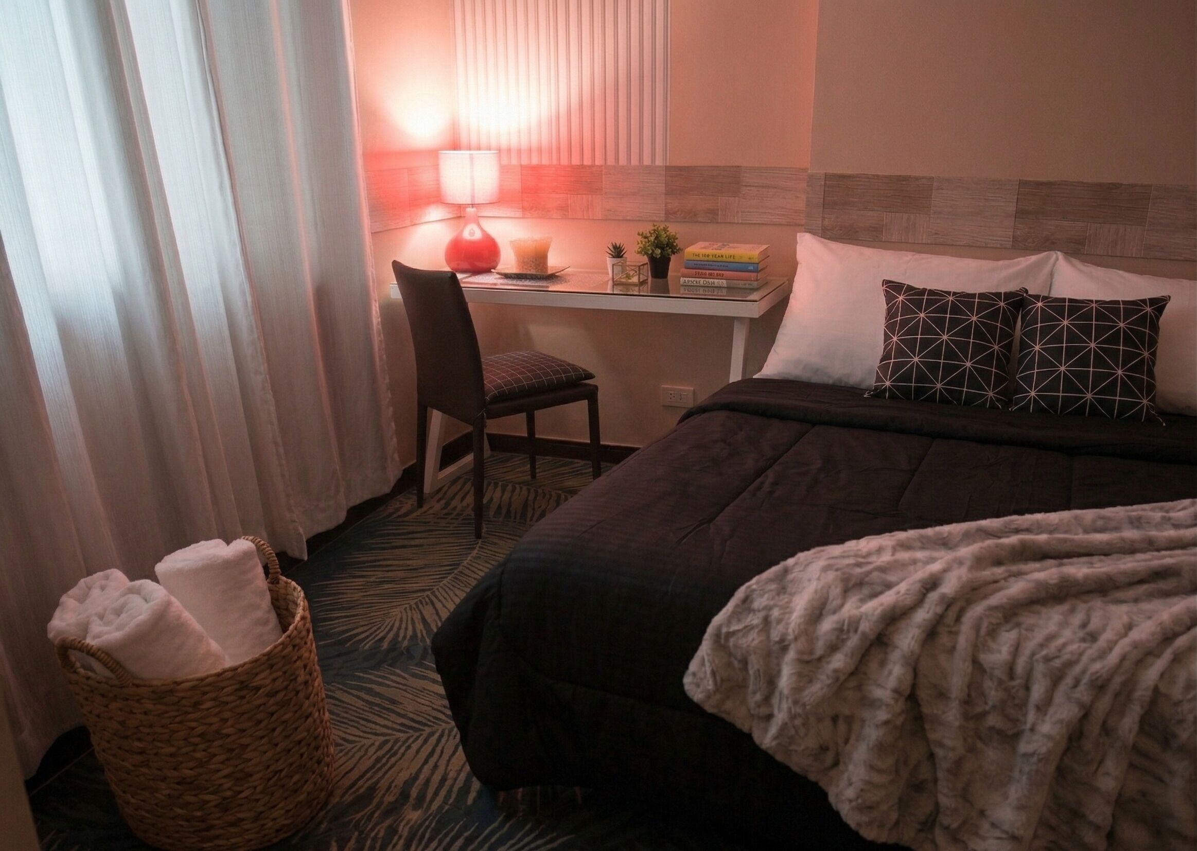 A hotel room with a bed, a desk with a chair, a lamp, a basket with towels, and a window with curtains.