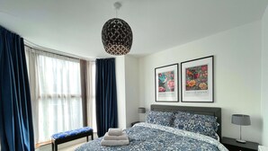 Quarto com uma cama grande, duas obras de arte emolduradas, um banco e um candeeiro suspenso.