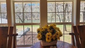 Une pièce avec une table en bois, un vase de tournesols et trois fenêtres donnant sur un paysage naturel.