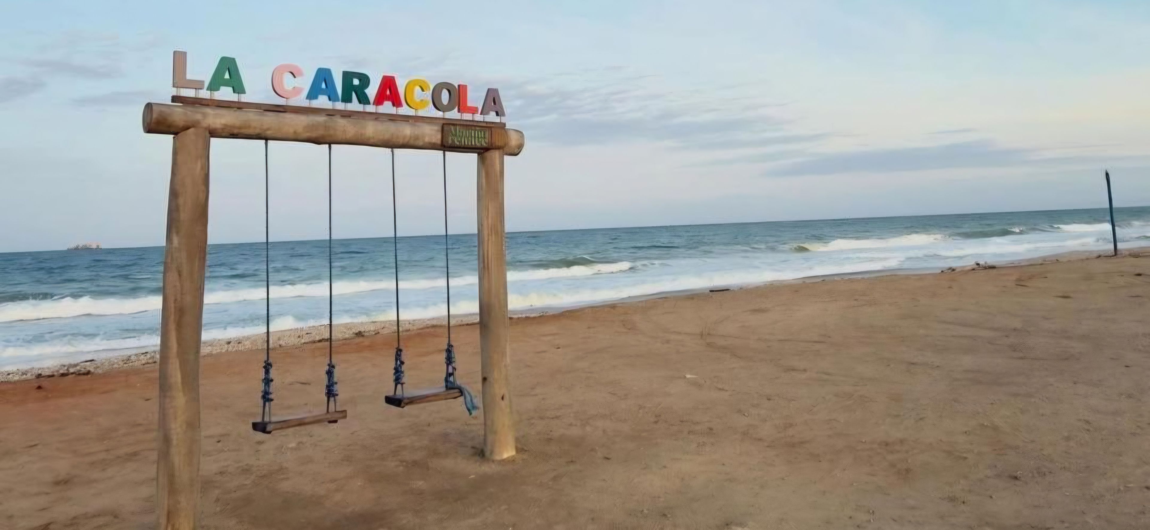 Een houten schommelset met het bord 'LA CARACOLA' op een strand, met de oceaan op de achtergrond.