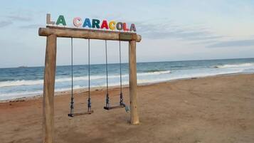 Een houten schommelset met het bord 'LA CARACOLA' op een strand, met de oceaan op de achtergrond.