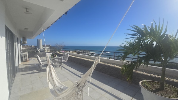 A balcony with a hammock, outdoor furniture, a potted plant, and a view of the sea and buildings.