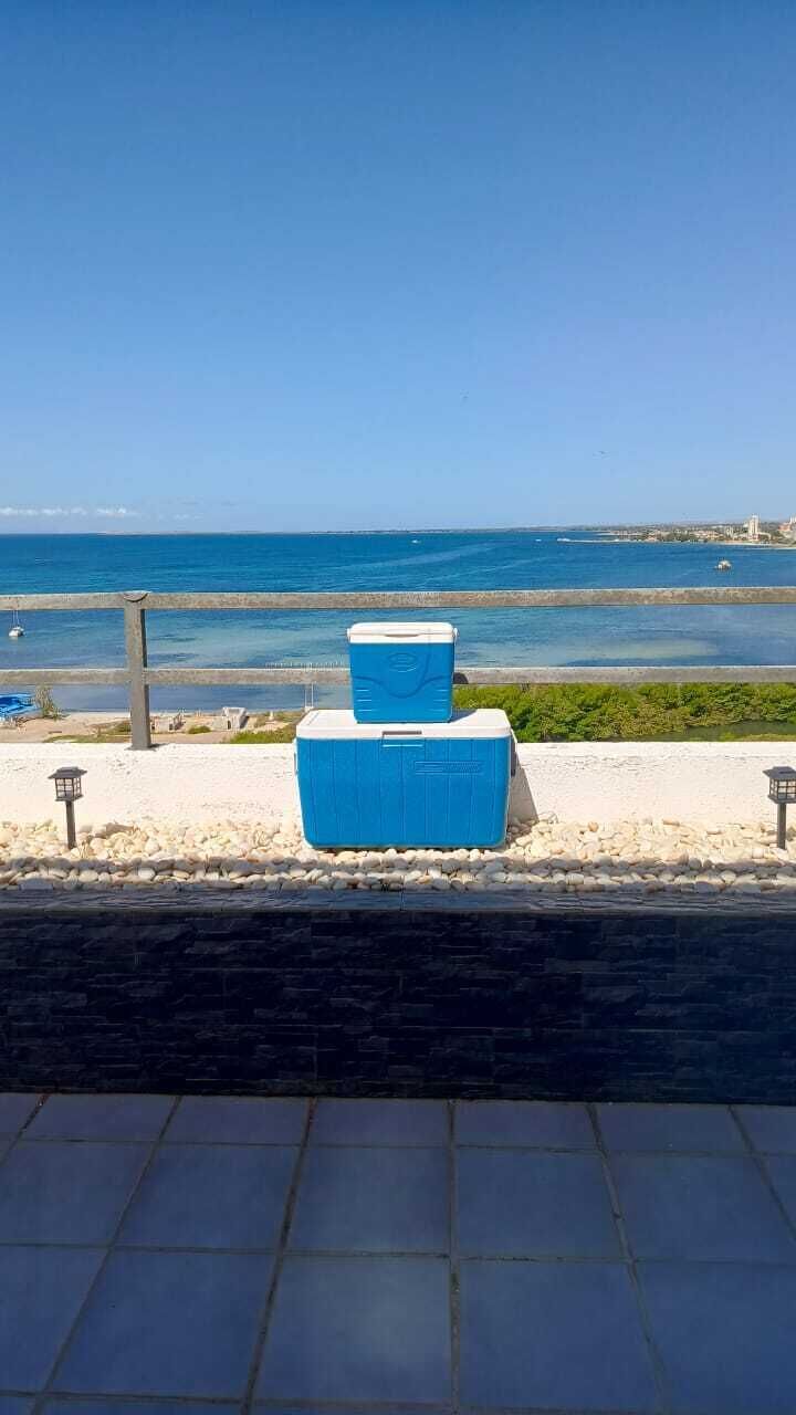 Une vue d’une plage avec un ciel dégagé, une étendue d’eau et deux glacières bleues posées sur une surface carrelée.