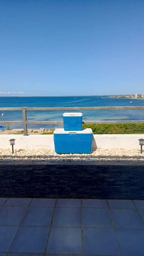 A view of a beach with a clear sky, a body of water, and two blue coolers on a tiled surface.