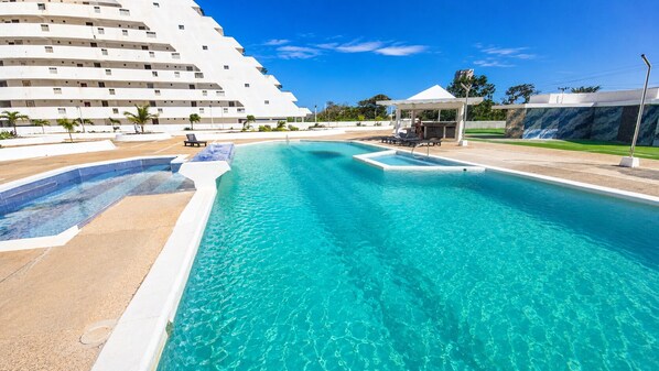 A large swimming pool with clear blue water, surrounded by a sandy area and a white building with multiple balconies.