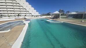 A swimming pool with a clear blue water, a white building with multiple floors, and a covered area with a roof.