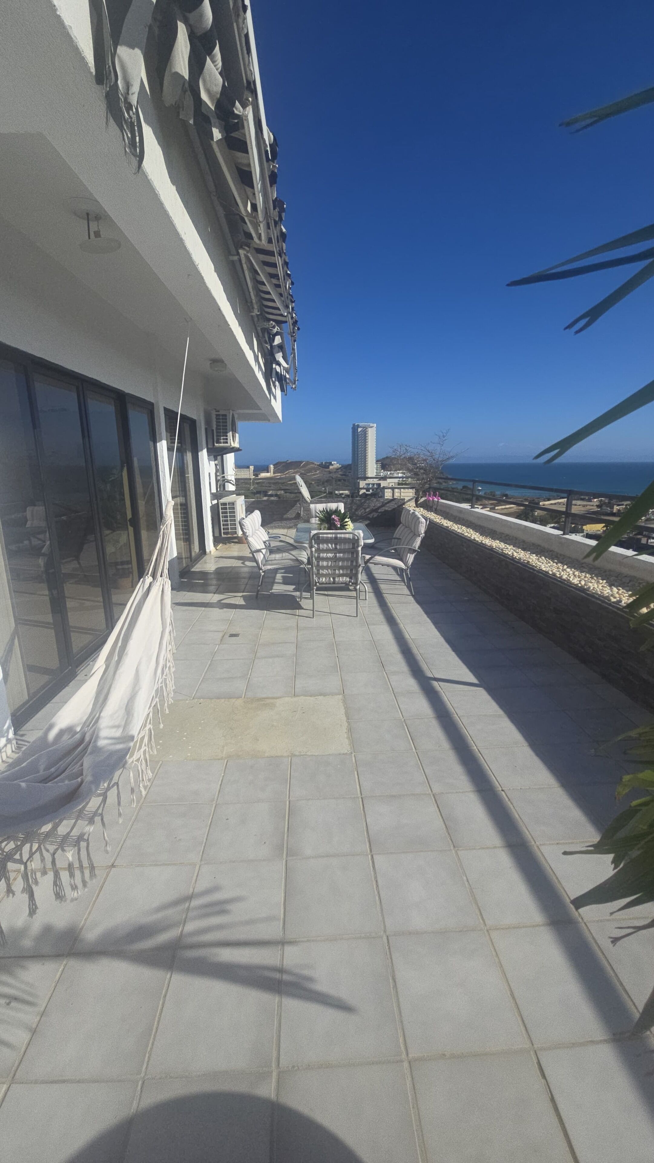 Una terraza en la azotea con mobiliario exterior, vistas al mar y un edificio con grandes ventanales.