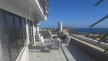 Una terraza en la azotea con mobiliario exterior, vistas al mar y un edificio con grandes ventanales.