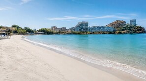 Une plage avec une eau cristalline, un rivage sablonneux et des bâtiments en arrière-plan.