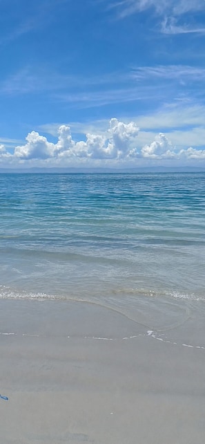 A beach with clear water, a sandy shore, and a blue sky with clouds.