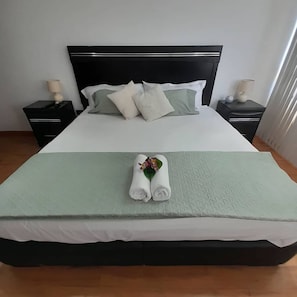 Una cama bien hecha con una colcha verde, almohadas blancas y una toalla enrollada con una flor. Hay dos mesitas de noche con lámparas.