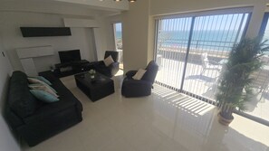 A modern living room with a sofa, armchair, and coffee table, a flat-screen TV, and a large window with a view of the ocean.