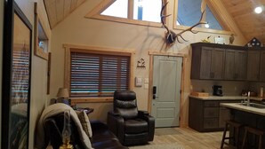 A cozy room with a leather sofa, a dining table, a kitchen area, and a large window with blinds. - The River Run Cabin (Ashton)