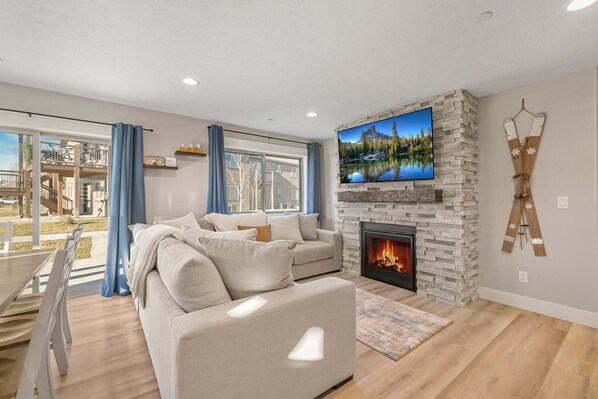 A modern living room with a fireplace, a television mounted on a stone wall, and a dining area with a table and chairs. - Victor Dreamweaver (Victor)