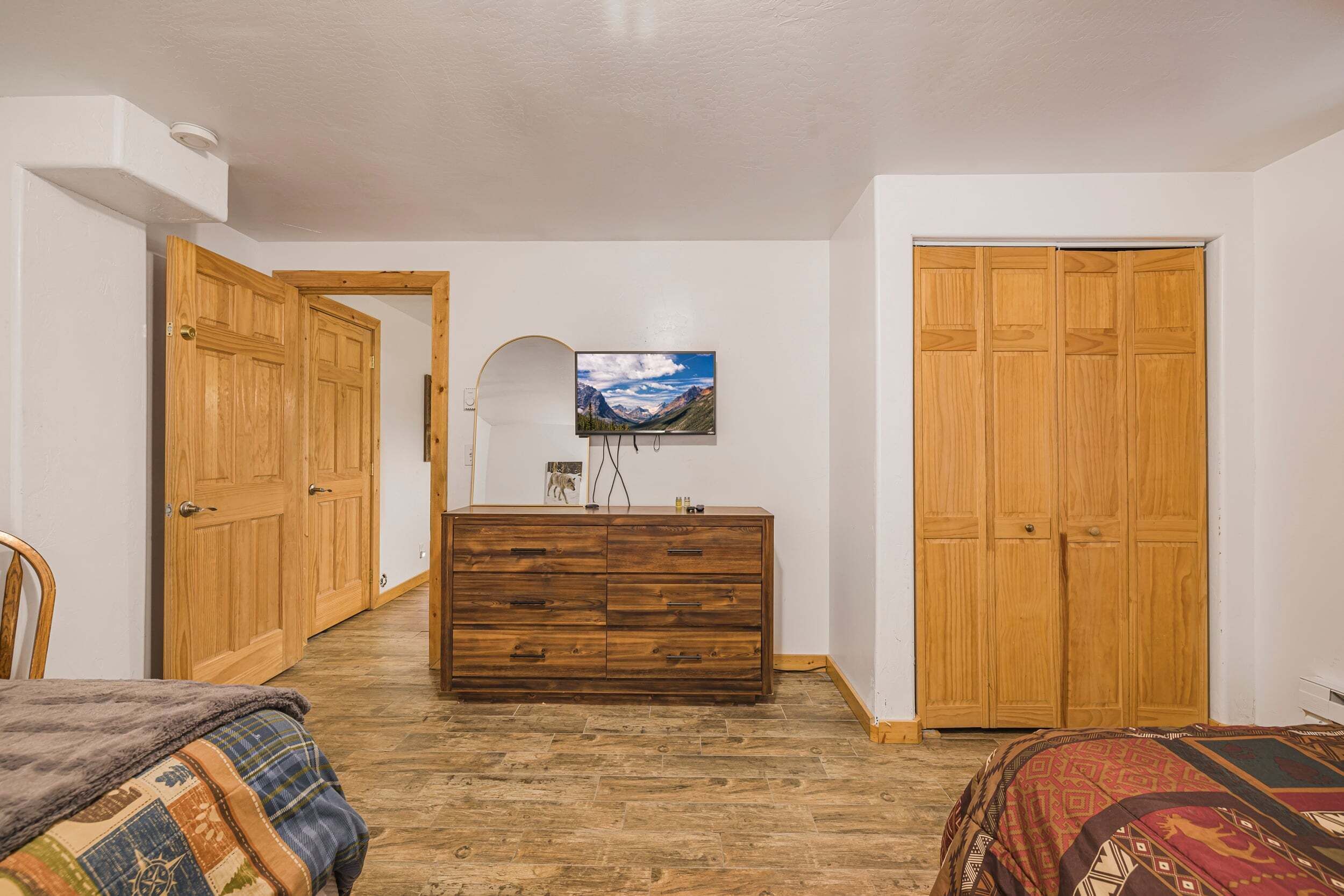 A bedroom with wooden wardrobes, a dresser, a television on the wall, and a patterned bedspread.