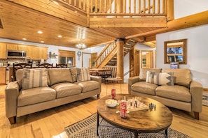 A spacious living area with two leather sofas, a wooden coffee table with snacks, a staircase, and a kitchen in the background.