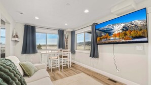 Un soggiorno moderno con una grande TV a schermo piatto, un'area pranzo con sedie, un divano con una coperta e vista sulle montagne dalle finestre.
