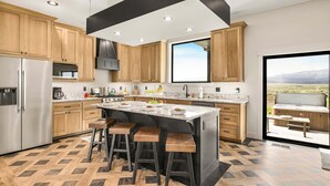 A modern kitchen with wooden cabinets, a large island with stools, a stainless steel refrigerator, and a view of a patio. - Checkmate (Island Park)