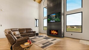 A modern living room with a leather sofa, a flat-screen TV, a fireplace, and large windows offering a view of the outdoors. - Checkmate (Island Park)