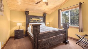 A bedroom with a wooden bed, a ceiling fan, a nightstand, a window with curtains, and a view of trees outside. - Jensen Pines (Island Park)