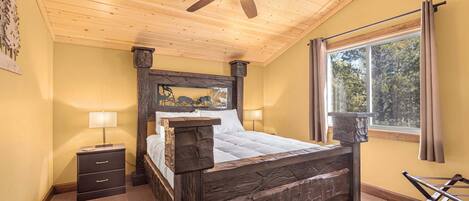 Un dormitorio con cama de madera, ventilador de techo, mesita de noche, ventana con cortinas y vistas a árboles.