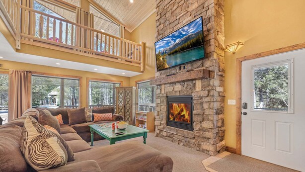 A spacious living room with a stone fireplace, a television mounted above it, and a balcony with wooden railings. - Jensen Pines (Island Park)